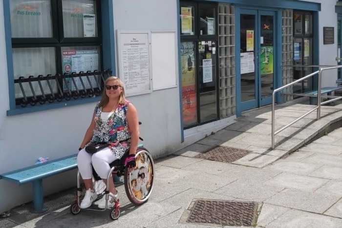 Disability activist Vicky Matthew - wheelchair user wearing sunglasses, a bright top with white trousers - outside a Bus &Eacute;ireann office which has a ramp leading to its door. 