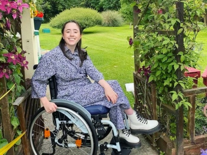 Niamh N&iacute; Hoireahbhaird, a wheelchair user wearing a grey patterned dress and cream Dr Martens boots, in her garden beside a climbing plant with pink flowers. 