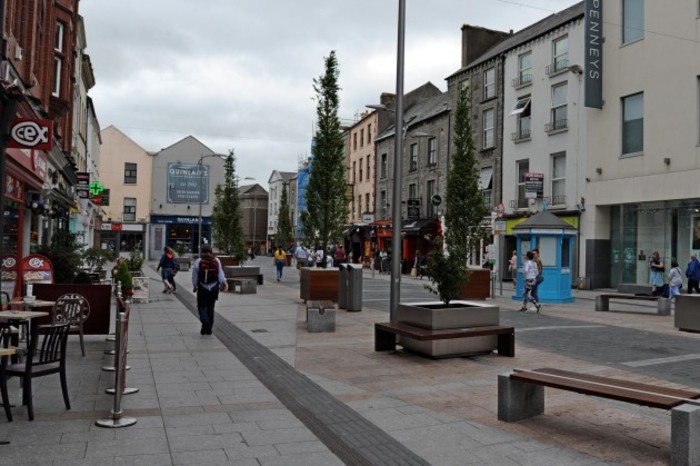 The pedestrian zone of the Mall in Tralee - a street lined with shops and cafes - which has no kerbs and is all on the same level, with some tactile paving beside a path that lies between the road section and seating areas. There are a number of planters with trees lining the road section.