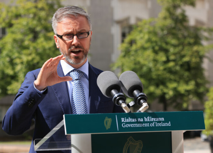 Minister Roderic O'Gorman - wearing a navy suit with light blue shirt and blue dotted tie - talking at a podium with two microphones on it. 