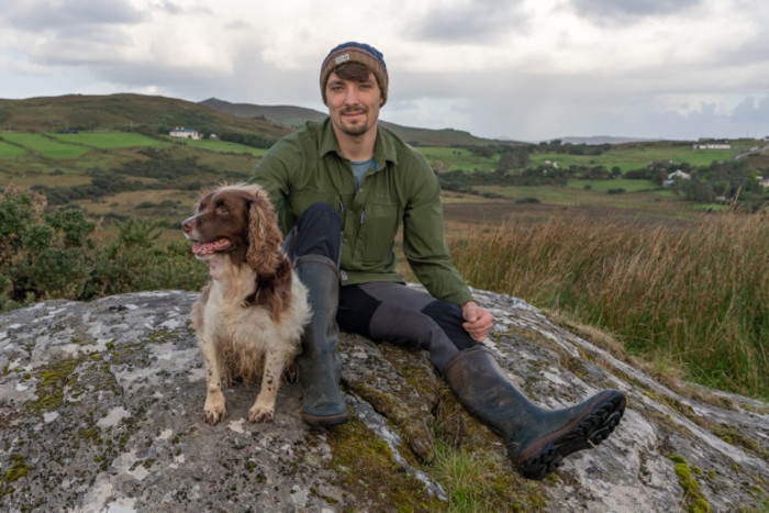Josh Mathews in warn, winter farm clothes sitting on top of a large rock on the family farm with Moose the dog in Co Mayo