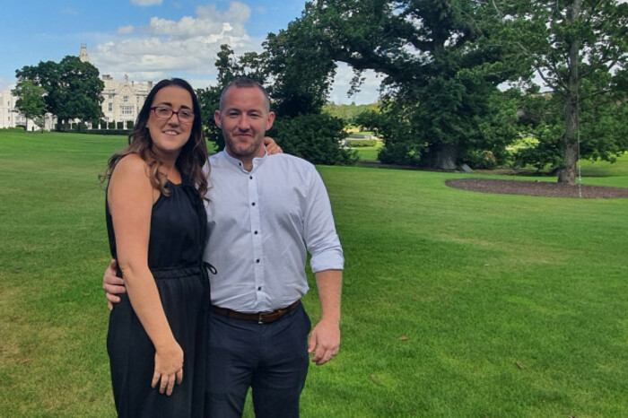 Rita - wearing a black dress - and Shane O'Meara - wearing a light blue shirt and trousers - standing on a lawn in a parkland with a building in the background.