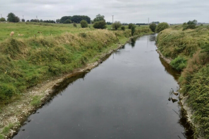 A straight section of river with banks with low grass and little other vegetation on both sides.