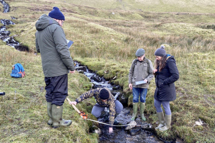A group of researchers wearing waterproofs including wellies standing in and beside a small stream. One researcher is hunkered down taking measurements. 