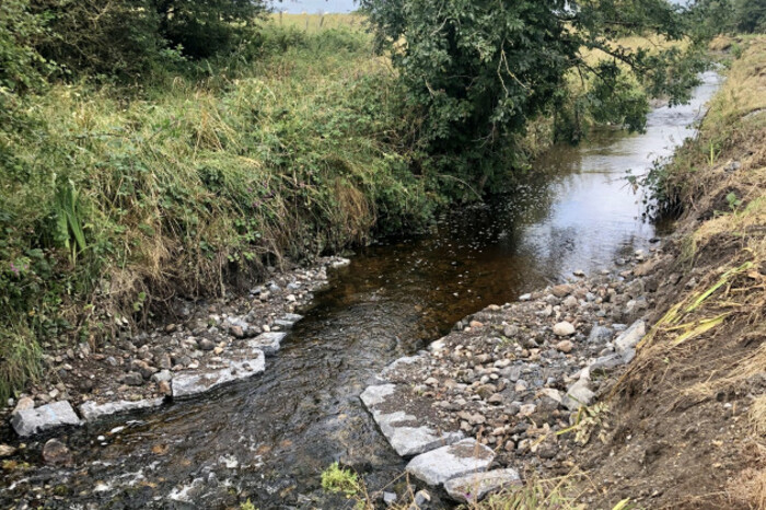 Large stones in a semi-circular line on both sides of the river, with gravel filled in behind these to narrow the river.