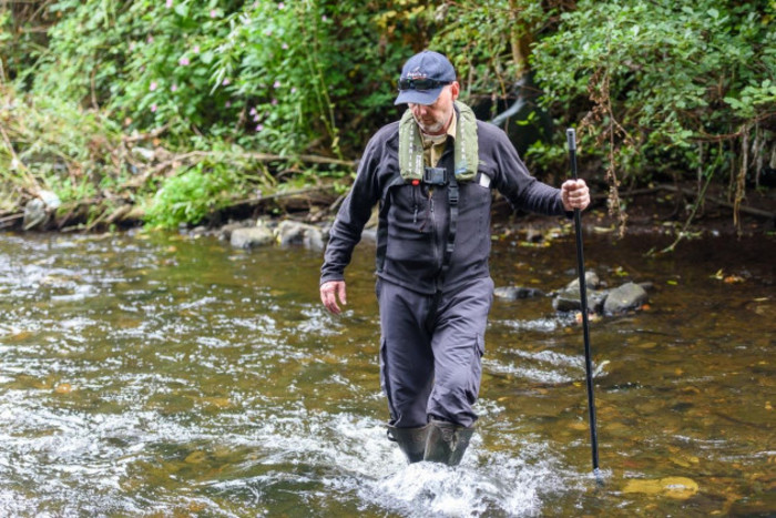 Declan Cooke wearing safety gear and wellies, while holding a large stick, walking through a river.