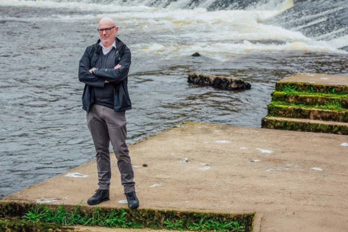 Will O'Connor wearing a jacket and trousers standing on a cement platform beside a river with a weir in the background.