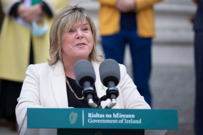 Minister Mary Butler wearing a white jacket, black top and necklace talking into microphones at a Government of Ireland podium. 
