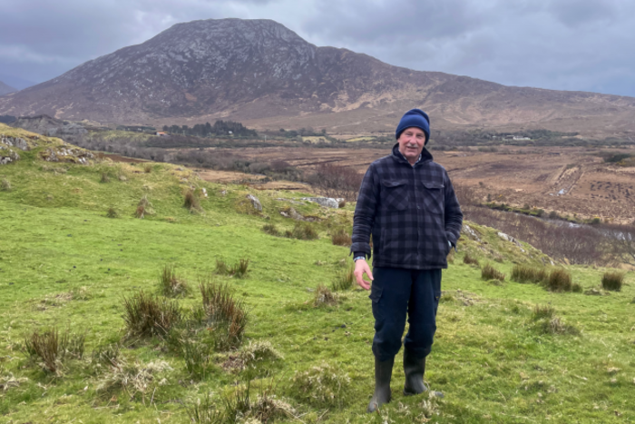 Tom Keane stands with a hat, jacket and wellies in a field with a river and mountain in the background.