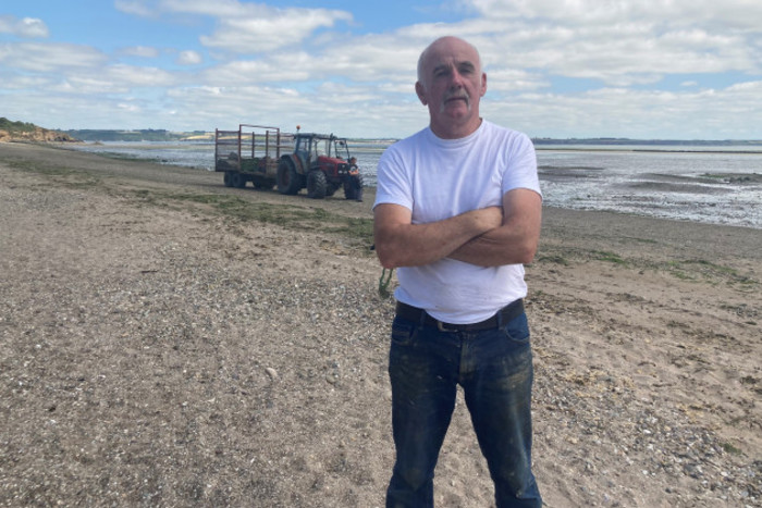 Pat Moran with his arms folded standing on a beach with a tractor and trailer behind near the water.