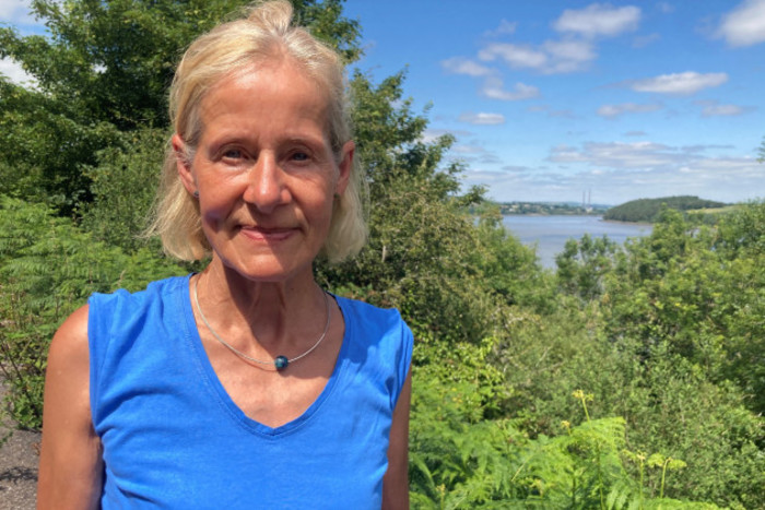 Karin Dubsky wearing a blue top and necklace standing in front of trees with the chimneys from the power plant visible in the distance. 