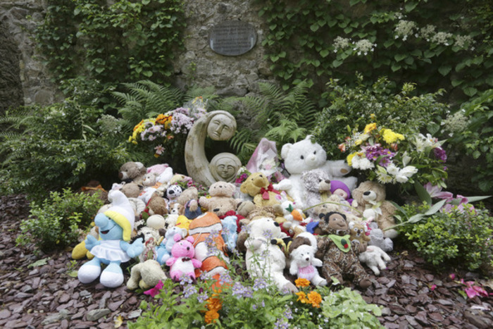 A large pile of stuffed animals including small teddy bears are placed beside a stone sculpture depicting a mother and child in a garden with a plague, ferns and other greenery.