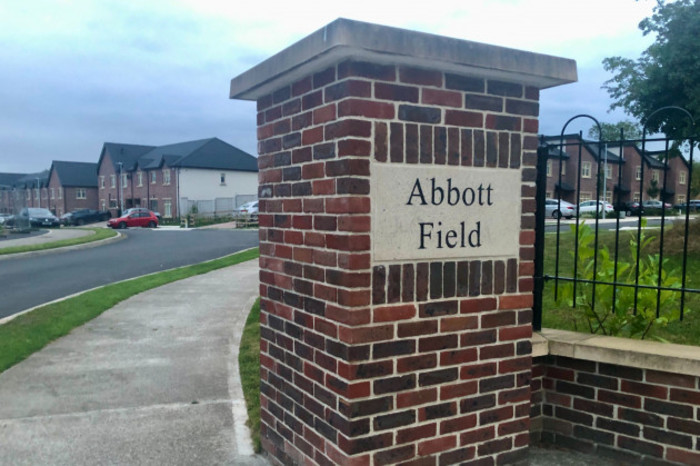  The entrance to Abbott Field with its sign integrated into a brick pillar. Two story houses with brick fronts and grey slate roofs can be seen in the background. 