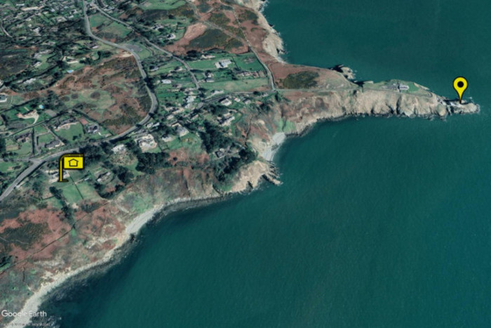 A satellite view of Howth head where the land and cliffs meet the sea along a rugged coastline. Roads and houses can be seen with a yellow marker on the left at a large housing complex with views over the sea. Another marker nearby is where Baily lighthouse stands out on a peninsula. 
