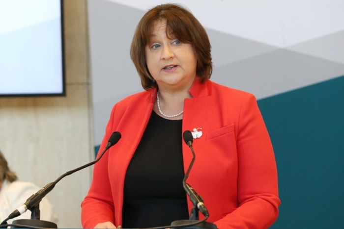 Minister Anne Rabbitte wearing a red jacket over a black top - speaking at a podium