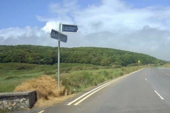 One of the three signs on the pole at the treated area points to Kilmurrin Cove. There are green fields and a forest in the background.