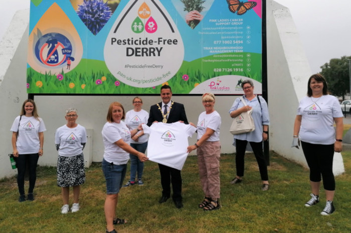 Seven women with Pesticide-Free Derry t-shirts are standing in front of a billboard which also says Pesticide-Free Derry. Two of the women are handing the then-mayor a t-shirt. 