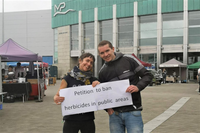 Virginia and James are holding a paper sign saying - Petition to ban herbicides in public places - and are standing in front of the shopping centre.