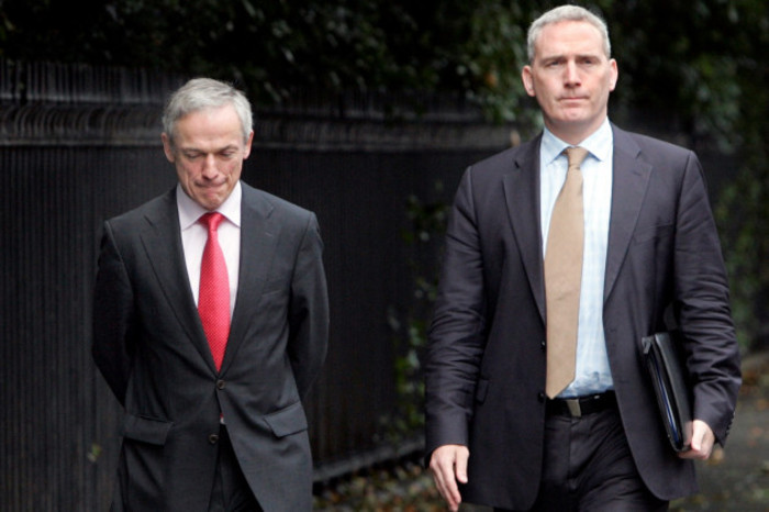 Richard Bruton - wearing a black suit with red tie - and Ciar&aacute;n Conlon - wearing a dark grey suit and pink tie - are walking beside each other on a Dublin street. Conlon is holding documents in one hand.