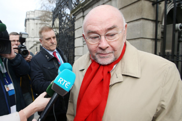 Ruairi Quinn wearing a beige coat and red scart outside the gates of Leinster House with an RT&Eacute; and other microphones being held towards him by reporters.