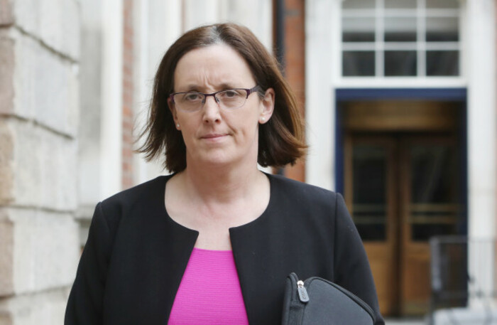 Helen Hall walking out of a stone building wearing a pink top and black jacket while holding a bag.