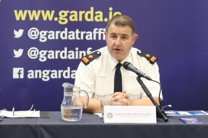 Michael Corbett at a conference speaking in a microphone with a sign in the background for the Garda website and social media addresses.