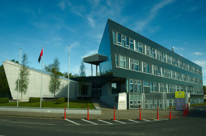 Dark grey four-story modern building with many white-framed windows. There are three flag poles outside it with a black and red flag hanging from one. 