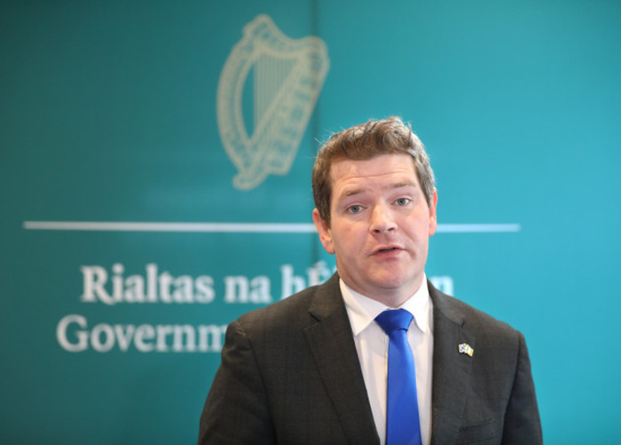 Peter Burke speaking - wearing a dark grey suit jacket and blue tie. The text - Rialtas nah&Eacute;ireann, Government of Ireland - is blurred in the background behind him.