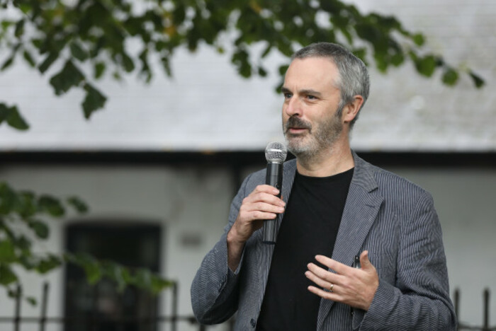 Fred Logue speaking outside while holding a mic. He is wearing a black top with a grey jacket.