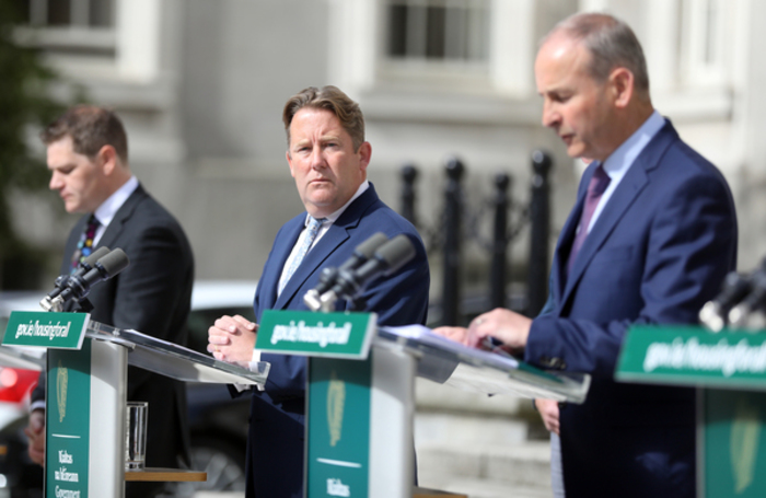 The two ministers and Taoiseach Martin are standing at podiums outside. Minister O'Brien is looking towards the Taoiseach while he speaks.