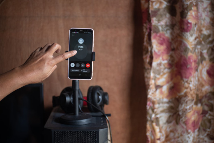 A phone is showing a call from &lsquo;Papa&rsquo; with a person with pointed finger about to touch the point. It is located in a room with a patterned curtain.