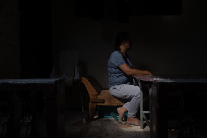 A woman sits at a desk in a darkened room to protect her identity. 