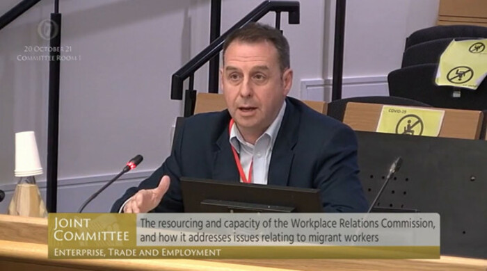 O'Brien wearing a white shirt, red lanyard and dark suit jacked sitting in the front of a number of rows of seats speaking into a microphone that is illuminated as being live.