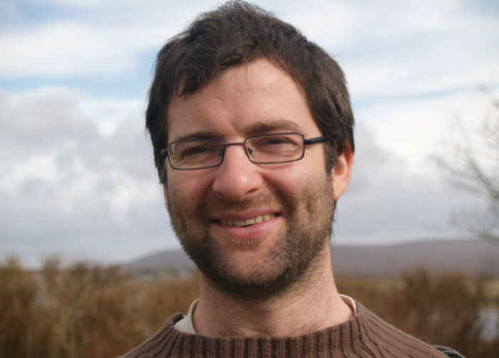 Photo of Eoin O'Leidhin, smiling in a brown jumper with trees and a cloudy sky in the background