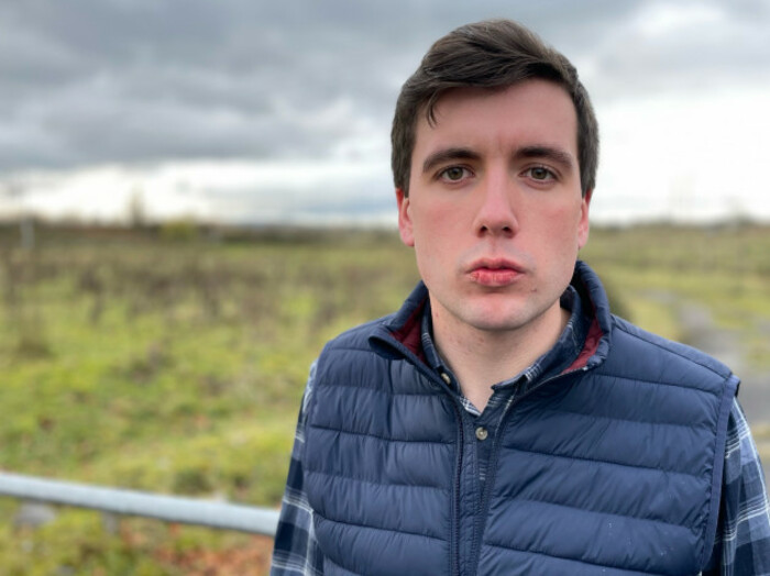 Tim Hannon of Futureproof Clare in a grey plaid shirt and navy windbreaker standing in front of a gate into a field.