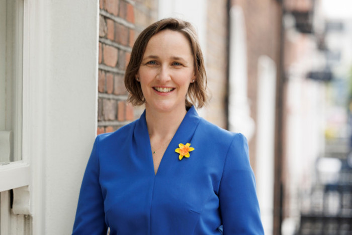 Amy Nolan wearing a blue jacket with a daffodil pinned to it, standing beside a red brick building.