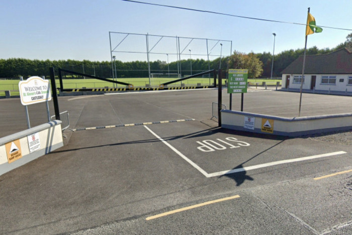 The entrance to the carpark which has a sign stating St Kieran's GAA Grounds Castledaly on the left side and a sign about the GAA jackpot on the right. The changing rooms can be seen on the right with a GAA pitch in the background. A green and gold flag flies at the entrance. 