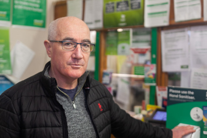 Clare Island native Padraic O'Malley in a dark grey jacket and glasses standing in front of the post office section of his shop