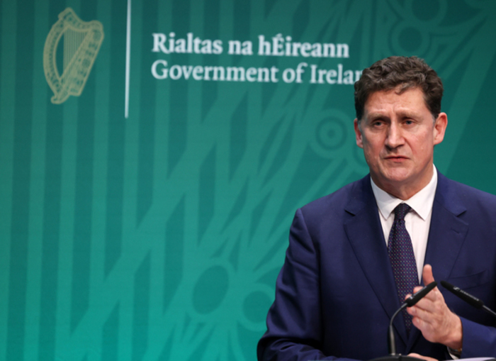 Eamon Ryan wearing a suit and tie speaking in front of a Rialtas na h&Eacute;ireann Government of Ireland sign.