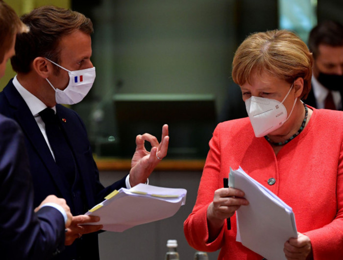 Angela Merkel and Emmanuel Macron - both wearing suits and face masks - holding documents while talking.