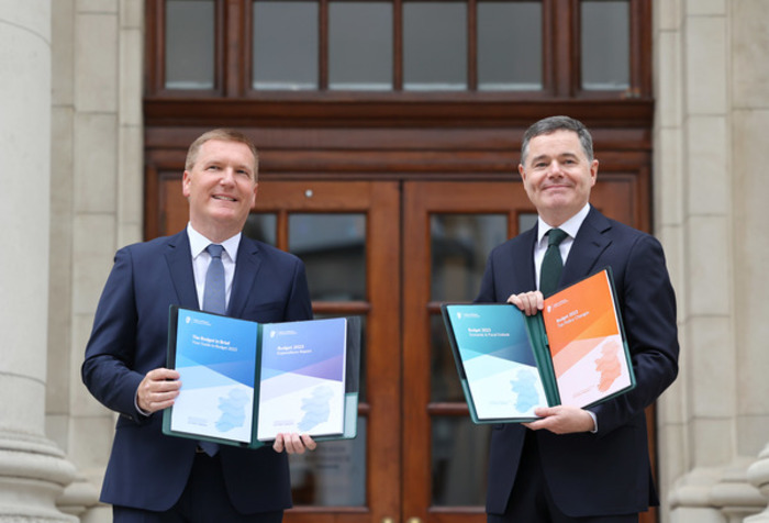 Ministers Michael McGrath and Paschal Donohoe standing outside a stone building with wooden doors - both holding Budget 2023 documents. 