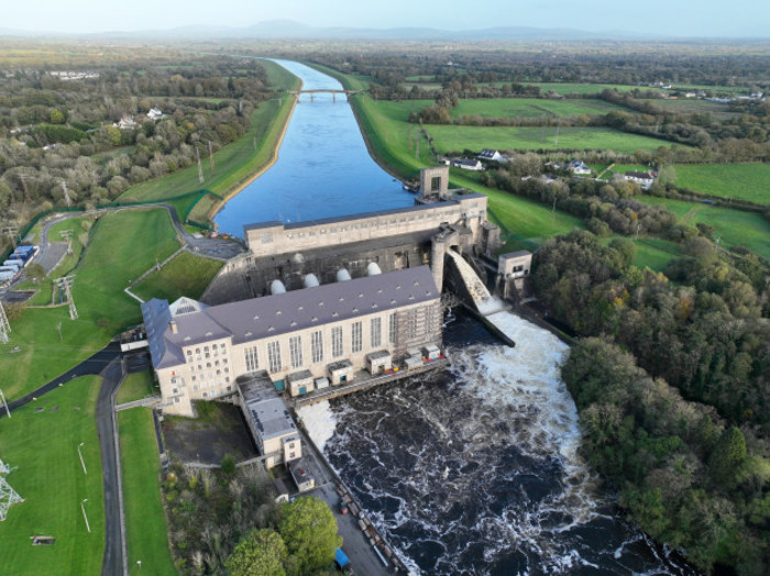 Ardnacrusha station consists of a large building with a grey roof in front with a dam which rises above it behind. Water is rushing down the spillway to the right of the dam. The river looks calmer behind the dam with turbulence visible in front. 