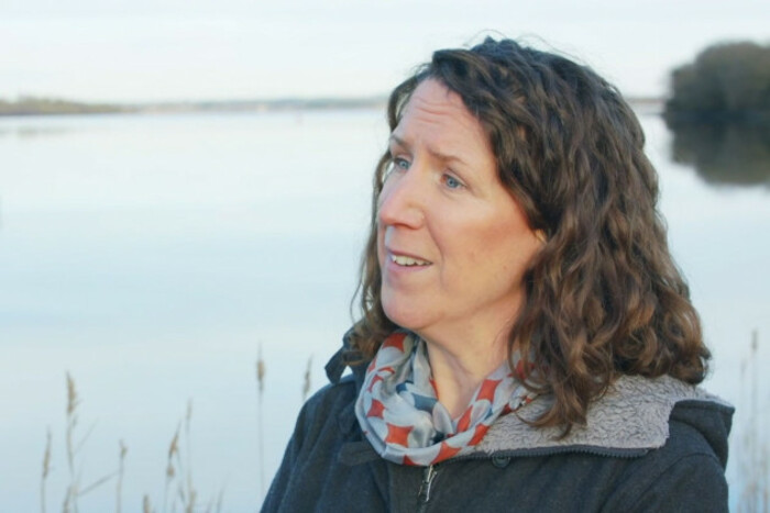 McGoff standing on the banks of a large body of water - looking towards an interviewer - wearing a warm coat and scarf. 