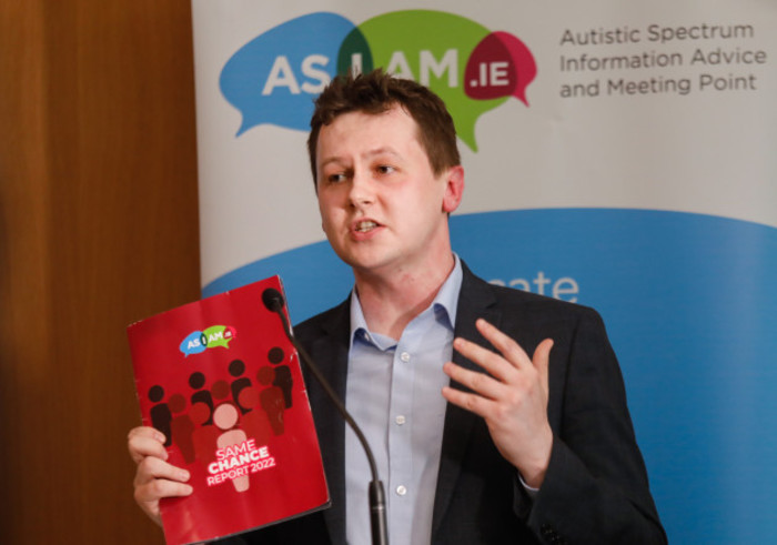 Adam Harris, the CEO of AsIAm, speaking from a podium in a suit with a report in his hands at an autism conference in 2022