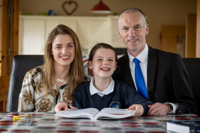 Cara Darmody dress in her navy school uniform smiling and holding an open book at her kitchen table with her parents to either side of her smiling