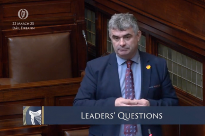 Richard O'Donoghue wearing a suit and tie speaking in the D&aacute;il chamber. D&aacute;il &Eacute;ireann 22 Mar 2022 and Leaders Questions are written on the screen.