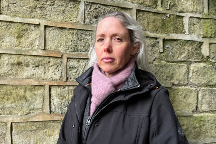 Leea Berry, a white woman with grey-blonde hair stands in front of a grey brick wall and looks at the camera with a serious expression. She is wearing a black coat and a pink scarf.