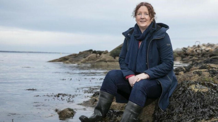 Dr R&oacute;is&iacute;n Nash smiling in a navy jacket sitting on a large rock beside the sea