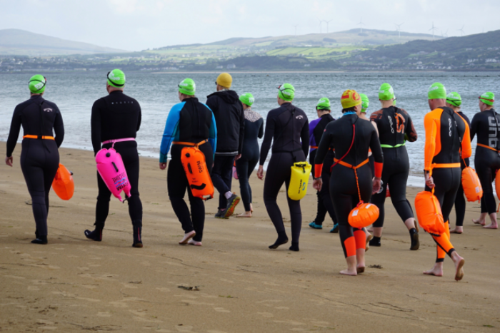 Swimmers wearing dark wetsuits and bright swimming caps walk away from the camera along the beach. Some have florescent buoys attached to their waists. In the background is Lough Swilly.