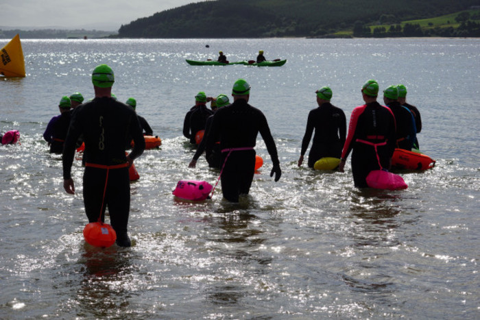 A group of swimmers wearing wet suits, green swim hats and pink, orange and yellow buoys walk into the sea. Two kayakers are monitoring the swim nearby. 